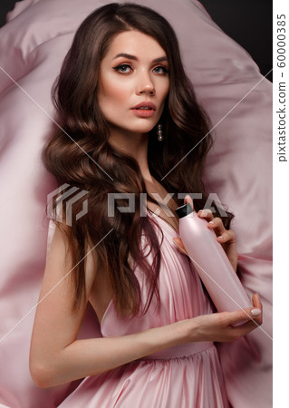Beautiful brunette woman with curls and classic make-up in a flying light pink dress with a bottle of shampoo in hands. Beauty face. Beautiful brunette woman with curls and classic make-up in a flying light pink dress with a bottle of shampoo in hands. Beauty face. 60000385