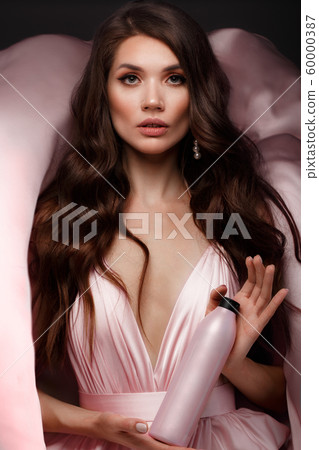 Beautiful brunette woman with curls and classic make-up in a flying light pink dress with a bottle of shampoo in hands. Beauty face. 60000387
