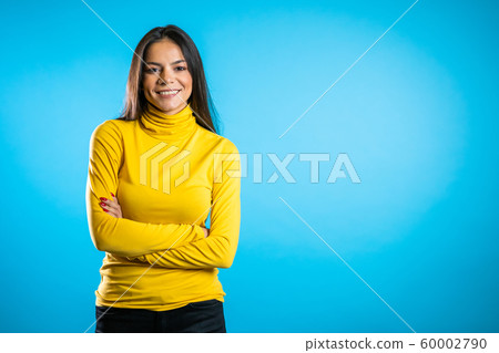 Copy space. Beautiful cheerful mixed race woman in yellow clothing smiling to camera over blue wall background. Cute hispanic girl's portrait. Copy space. Beautiful cheerful mixed race woman in yellow clothing smiling to camera over blue wall background. Cute hispanic girl's portrait. 60002790