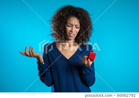 Pretty african-american young woman holding small jewelry box with marriage proposal on blue background. Disappointed upset girl, she doesn't like gift. Pretty african-american young woman holding small jewelry box with marriage proposal on blue background. Disappointed upset girl, she doesn't like gift. 60002792
