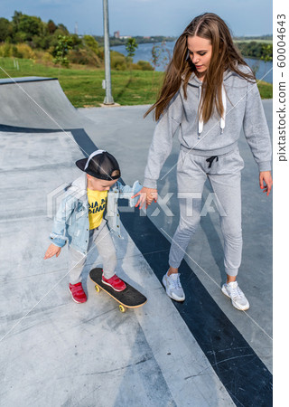 Young mother teaches to ride a skateboard, son of a little boy, summer day. Autumnal clothes, casual clothes. Help and support in parenting. Caring for children. Workout balance. 60004643