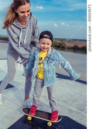 Young mother with little boy son, happy family learns to ride skateboard, summer outdoors, autumn day. Emotions of delight, joy of happiness and fun. Caring support assistance in training, parenting. 60004671