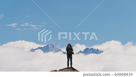 a man with backpack standing on mountain top, with snow mountains over clouds 60008434