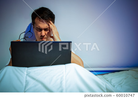 headache, tense young asian man working on laptop computer in bed at night 60010747