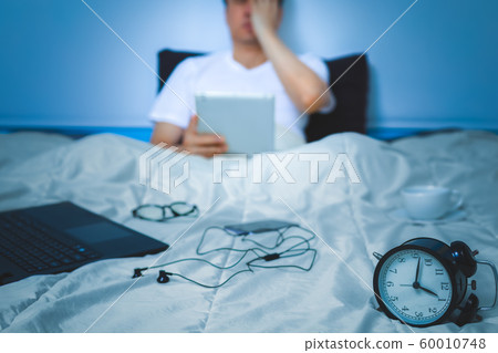 sleepless, headache, tense young asian man working on tablet pc, laptop computer and smartphone in bed at night 60010748