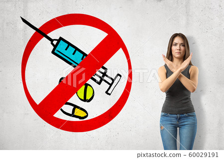 Serious young girl with hands crossed, standing against wall with picture of syringe and some meds enclosed in red prohibition sign. Serious young girl with hands crossed, standing against wall with picture of syringe and some meds enclosed in red prohibition sign. 60029191