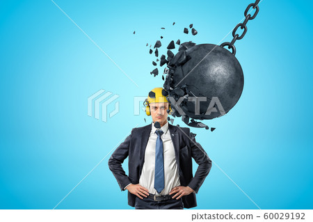 Businessman wearing yellow safety helmet and metal chained ball breaking into pieces on blue background 60029192