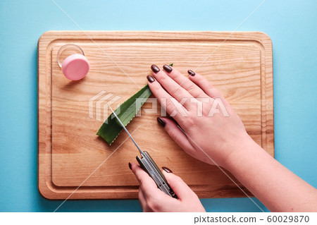 Cutting aloe vera slices with a knife next to a open empty box with pink cap, blue background, top view, woman hands 60029870