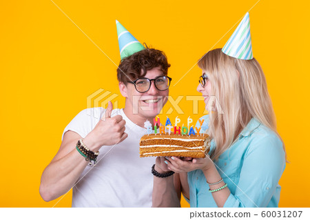 Young cheerful students charming girl and nice guy in greeting paper caps holding a cake. Concept of congratulations on the birthday and anniversary 60031207