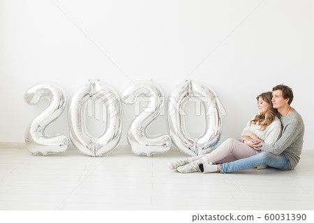 Holidays, festive and party concept - Happy loving couple sitting on a floor near silver 2020 balloons on white background. New Year celebration 60031390