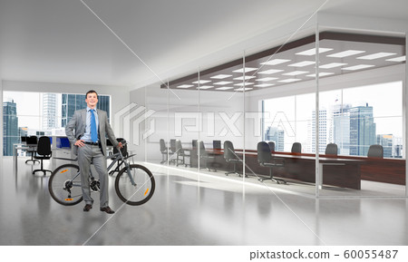 Smiling man in suit standing with bike in office Smiling man in suit standing with bike in office 60055487