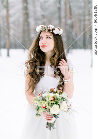 Winter wedding portrait. Portrait of beautiful bride with floral and cotton wreath on the head, holding bouquet, wearing a white dress against the background of the winter forest in snowy weather 60061286