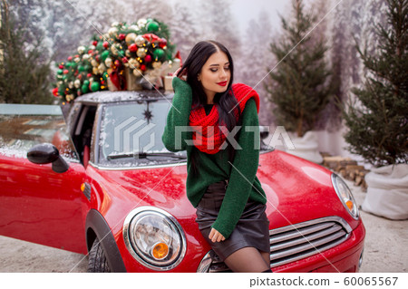 Pretty young girl is wearing red scarf sitting on red car with decorated xmas tree on the roof, holiday concept. 60065567