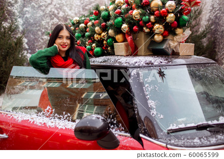 Pretty young girl is standing near red car with decorated xmas tree on the roof, holiday concept. Pretty young girl is standing near red car with decorated xmas tree on the roof, holiday concept. 60065599