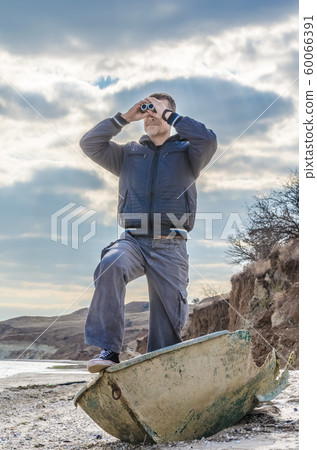 man in black stands in a broken boat on the beach and carefully looks through binoculars man in black stands in a broken boat on the beach and carefully looks through binoculars 60066391