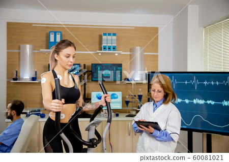 Female athlete running tests on the stepper and doctor making notes in tablet 60081021