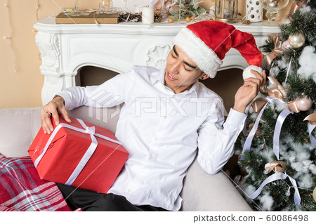 Young man in santa's hat sitting on couch near Young man in santa's hat sitting on couch near 60086498