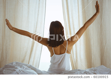 Woman stretching hands in bed after wake up, sun flare . Brunette entering a day happy and relaxed after good night sleep and back view. Concept of a new day and joyful weekend Woman stretching hands in bed after wake up, sun flare . Brunette entering a day happy and relaxed after good night sleep and back view. Concept of a new day and joyful weekend 60100915