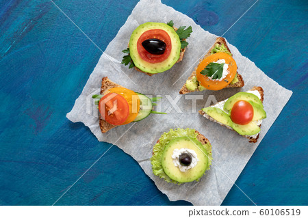 Vegetarian sandwiches with tomatoes, avocado, cucumber, green salad, parsley, cheese and sun-dried olives on parchment paper piece on blue background. Top view, flat lay, copy space. Vegetarian sandwiches with tomatoes, avocado, cucumber, green salad, parsley, cheese and sun-dried olives on parchment paper piece on blue background. Top view, flat lay, copy space. 60106519