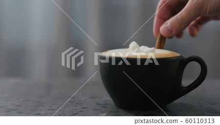 adding cinnamon stick into cappuccino in black cup on terrazzo countertop 60110313