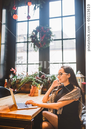 Beautiful young brunette woman inside cafe with christmas interior at wooden table by the window talking on phone and looking the laptop.In winter,she is dressed in a gray knitted sweater and glasses Beautiful young brunette woman inside cafe with christmas interior at wooden table by the window talking on phone and looking the laptop.In winter,she is dressed in a gray knitted sweater and glasses 60113619