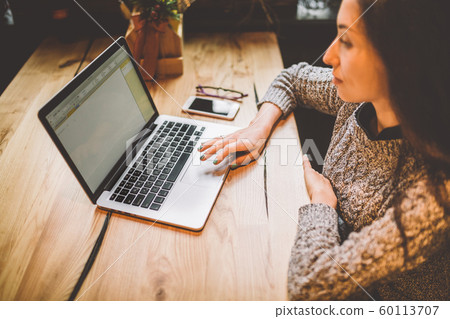 Close-up of a young girl's hand in a gray sweater use laptop technology on a wooden table in a cafe. Near the mobile phone and glasses. The concept of remote work business lady. Close-up of a young girl's hand in a gray sweater use laptop technology on a wooden table in a cafe. Near the mobile phone and glasses. The concept of remote work business lady. 60113707