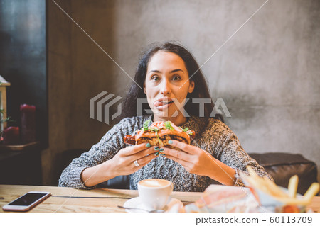 Dark-skinned brunette woman funny eating a burger in a cafe in a sweater. Emotion bulging eyes and tongue out Dark-skinned brunette woman funny eating a burger in a cafe in a sweater. Emotion bulging eyes and tongue out 60113709
