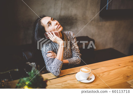 beautiful young brunette girl talking on mobile phone at wooden table near window and drinking coffee in cafe decorated with Christmas decor. Dressed in a gray knitted wool sweater beautiful young brunette girl talking on mobile phone at wooden table near window and drinking coffee in cafe decorated with Christmas decor. Dressed in a gray knitted wool sweater 60113718