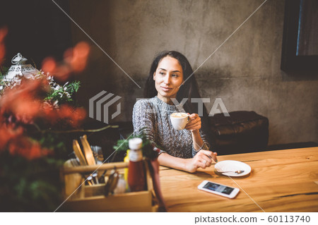 beautiful young girl drinks coffee from a white cup, next to her cell phone in a cafe decorated with Christmas decor. Dressed in a gray knitted wool sweater beautiful young girl drinks coffee from a white cup, next to her cell phone in a cafe decorated with Christmas decor. Dressed in a gray knitted wool sweater 60113740