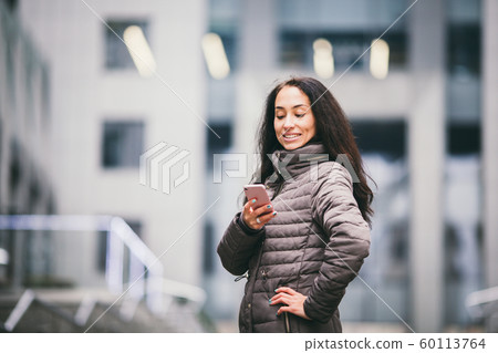 Business lady. Beautiful caucasian brunette woman uses technology mobile phone pink against background of the plastic facade of the window. The girl is calling, talking on the phone. Theme Business 60113764