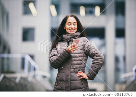 Business lady. Beautiful caucasian brunette woman uses technology mobile phone pink against background of the plastic facade of the window. The girl is calling, talking on the phone. Theme Business 60113765