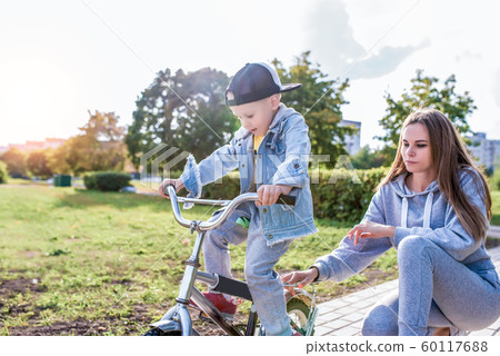 Mom woman teaches riding bike, little boy son 3-5 years old, training rides, in summer city, in autumn in park, casual clothes. Caring help and support in learning first lessons, balance. Mom woman teaches riding bike, little boy son 3-5 years old, training rides, in summer city, in autumn in park, casual clothes. Caring help and support in learning first lessons, balance. 60117688