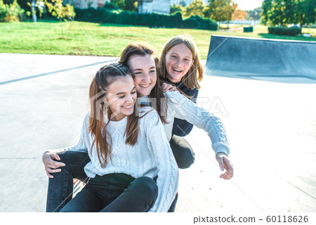 Three girls schoolgirls teenagers 13-15 years old, autumn day summer city, skateboarding relaxing after school on vacation. Emotions joy are fun enjoyment, happy are smiling laughing, casual clothes 60118626