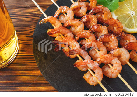 Shrimp fried on skewers with arugula, lemon on a black slate dish and beer in a glass on a wooden table 60120418