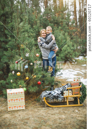 Pretty young mother holding her son standing near the decorated Christmas tree in winter forest outdoors. Wooden sledge and present boxes under the tree 60120717