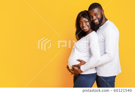 Pregnant afro woman and her husband embracing over yellow background Pregnant afro woman and her husband embracing over yellow background 60121540