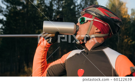 Training an athlete on the roller skaters. Biathlon ride on the roller skis with ski poles, in the helmet. Autumn workout. Roller sport. Athlete drinks water from flask. 60122639