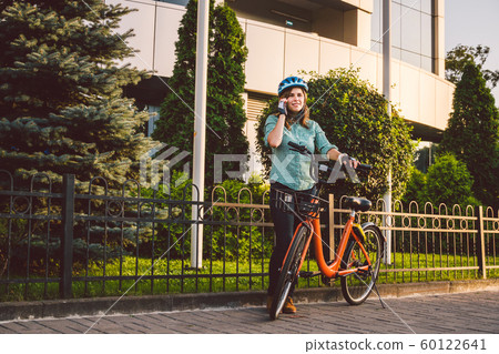 Portrait Of A Cheerful Girl Holding Mobile Phone. Happy smiling student using bike sharing app on smart phone outdoor. City lifestyle stylish hipster girl with bike using smartphone app in street Portrait Of A Cheerful Girl Holding Mobile Phone. Happy smiling student using bike sharing app on smart phone outdoor. City lifestyle stylish hipster girl with bike using smartphone app in street 60122641