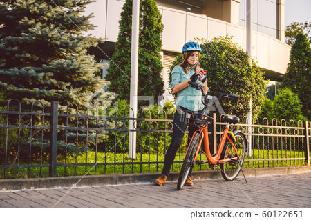 Portrait Of A Cheerful Girl Holding Mobile Phone. Happy smiling student using bike sharing app on smart phone outdoor. City lifestyle stylish hipster girl with bike using smartphone app in street Portrait Of A Cheerful Girl Holding Mobile Phone. Happy smiling student using bike sharing app on smart phone outdoor. City lifestyle stylish hipster girl with bike using smartphone app in street 60122651
