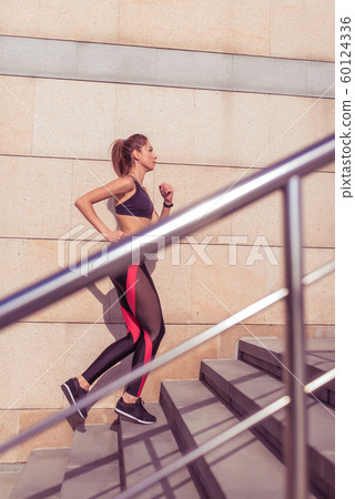 Young and athletic woman, runs up stairs, in summer on jogging in city, side view, sportswear. Workout athletics, day and morning. Active fitness lifestyle. 60124336