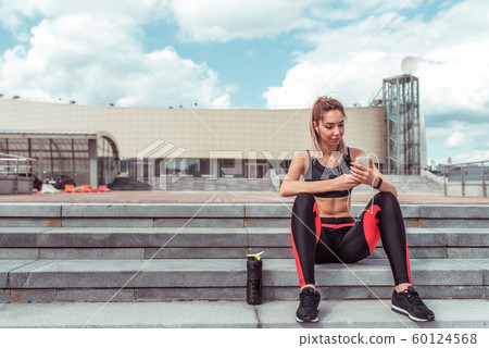 Girl athlete sits stairs rest workout, summer day city. Reads writes message on Internet, smartphone listens music headphones. Enjoys listens podcast lecture. Active lifestyle fitness, stylish woman. 60124568