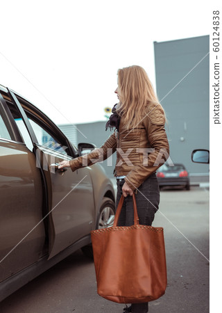 woman in parking lot of shopping center opens car door, in her hand is bag with groceries and purchases, brown leather jacket and bag, crossover car. Autumn in city. 60124838