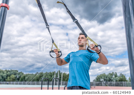 Athletic man exercises with straps loops trx, pulling up and training chest muscles, workout in summer in city, active lifestyle, modern fitness workout. 60124949