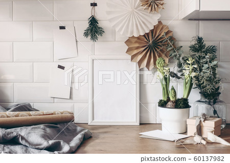 Christmas still life. Blank wooden picture frame and greeting cards mockup with paper stars, eucalyptus, potted hyacinth flowers and gift wrapping papers. White tiles background. Winter composition. 60137982