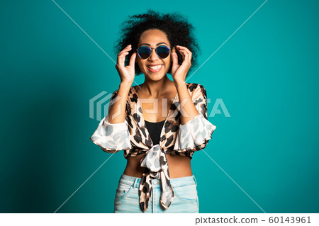 Pretty african woman with afro hair in leopard top and sunglasses smiling to camera over blue wall background. Cute mixed race girl's portrait. Pretty african woman with afro hair in leopard top and sunglasses smiling to camera over blue wall background. Cute mixed race girl's portrait. 60143961