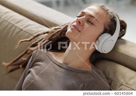 Relaxed girl with headphones smiling while listening to calming music on couch 60145804