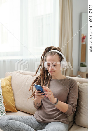 Young female with dreadlocks scrolling in smartphone while sitting on couch Young female with dreadlocks scrolling in smartphone while sitting on couch 60147403
