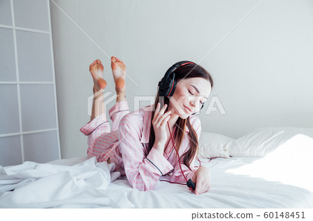 brunette in pink Pajamas listening to music with headphones on the bed 60148451