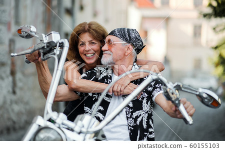 A cheerful senior couple travellers with motorbike in town. 60155103