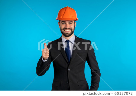 Positive young engineer businessman in construction helmet smiles to camera. Man showing thumbs up sign over blue background. Winner. Success. Body language. 60157072